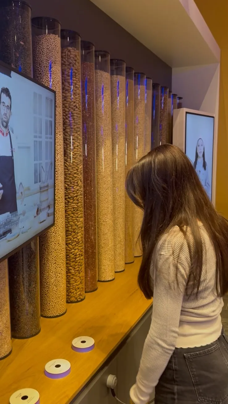 Diyetisyen Elif Beyza Kaya Sarper, Çatalhöyük sergisinde tahıl ve baklagil tüplerinin önünde; ekranda o dönemin yemeklerini anlatan bir şef